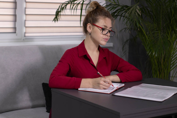 blonde girl in a red shirt and red glasses works - writes in a notebook. In a gray office on a gray sofa at a gray table - Photo, Image