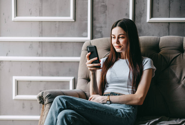 Beautiful young smiling caucasian woman resting on a sofa and using smartphone. Chatting with friends, browsing information. High quality photo - 写真・画像