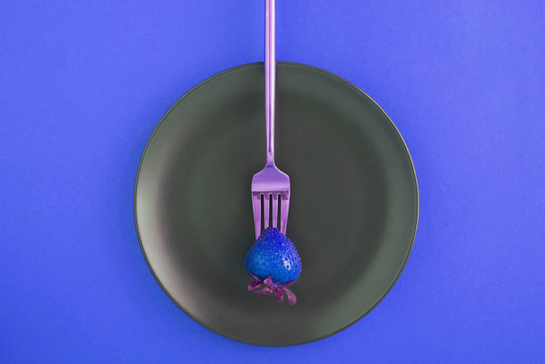 Una fresa azul empalada en un tenedor de oro en el plato negro sobre el fondo azul. Vista superior
. - Foto, imagen