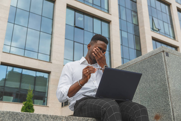 Feeling exhausted. Frustrated young black african man keeping eyes closed and looking tired from extra working on laptop - 写真・画像
