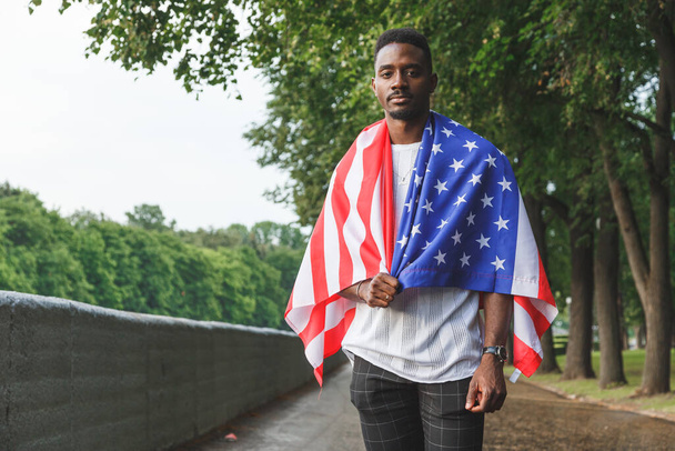 Όμορφος Αφρο-Αμερικανός με αμερικανική σημαία στους ώμους του κοιτάζει σοβαρά την κάμερα, στέκεται έξω. Καλοκαίρι. - Φωτογραφία, εικόνα