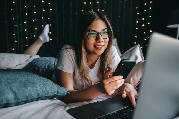 Beautiful young smiling caucasian woman resting on a bed at home and using laptop computer and smartphone. Chatting with friends, shopping online. High quality photo - Foto, Imagen