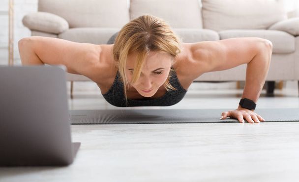 Sportnő edzés otthon. Izmos felnőtt nő fitness tracker csinál push-up, közel laptop a nappaliban belső - Fotó, kép
