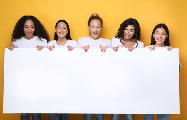 Opgewonden meisjes houden lege poster voor tekst, gele achtergrond, Mockup - Foto, afbeelding
