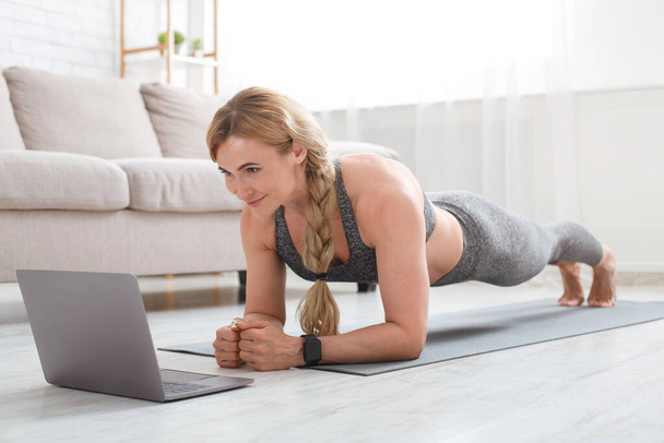 Fitness egzersizi. Spor giysili bir kadın paspasa tahta örüyor ve dizüstü bilgisayara bakıyor. - Fotoğraf, Görsel