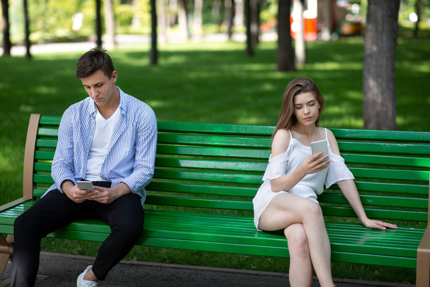 Gadget adicción y problemas de relación. Pareja joven con teléfonos inteligentes sentados en el banco en el parque, ignorándose mutuamente
 - Foto, imagen