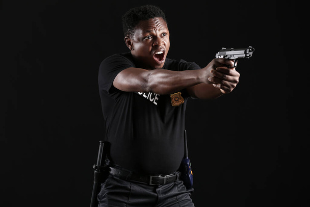 Aggressive African-American police officer with gun on dark background - 写真・画像
