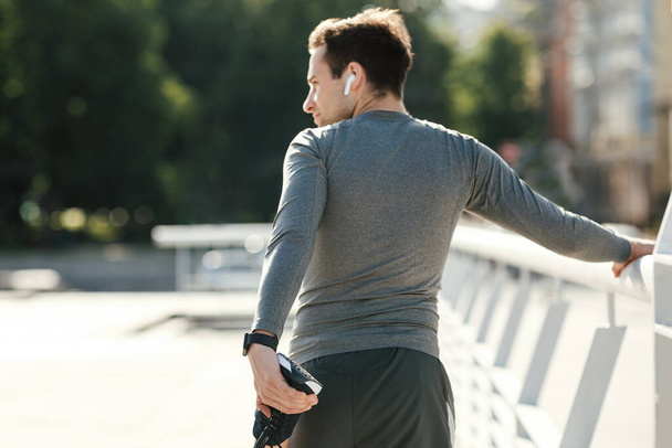 Estirar las piernas para el entrenamiento. El hombre con auriculares inalámbricos modernos hace ejercicios para correr
 - Foto, imagen