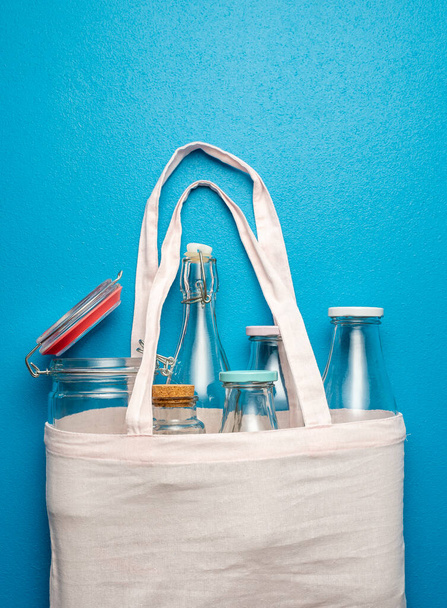 Вид зверху з порожніми скляними пляшками і баночками у бавовняній сумці. Пляшки без пластику в продуктовій сумці на синьому фоні
. - Фото, зображення