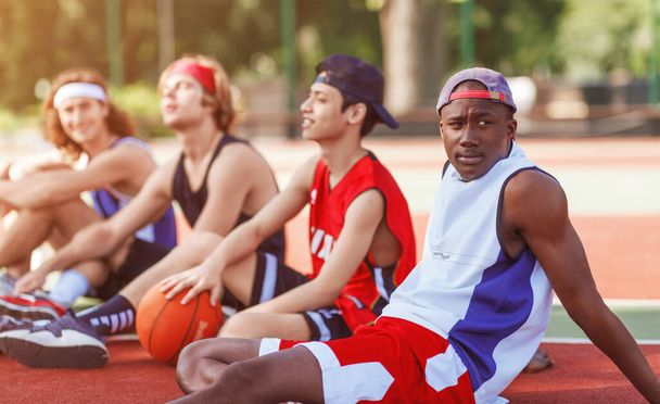 Μαύρος μπασκετμπολίστας με την ποικιλόμορφη ομάδα του να ξεκουράζεται στο υπαίθριο γήπεδο μετά από ένα δύσκολο παιχνίδι, ελεύθερος χώρος - Φωτογραφία, εικόνα