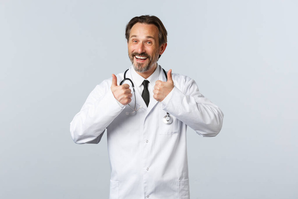 Covid-19 、コロナウイルスの発生、医療従事者およびパンデミックの概念。白衣を着た熱狂的な喜ぶ男性医師が承認に親指アップを示しています.クリニックの医師はプロモーションをお勧めします - 写真・画像