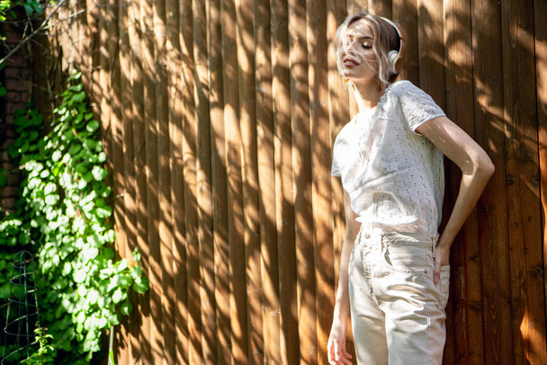 Портрет красивой девушки в беспроводных наушниках, стоящей напротив деревянного забора на заднем дворе в солнечный день
. - Фото, изображение