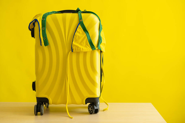 A candid mini-bikini of the coloring of the Brazilian flag hangs on a suitcase on a yellow background. The concept of a summer beach holiday. A woman is going on a trip to the sea. - Photo, image