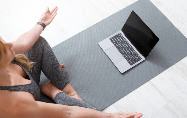 Allenati a casa online. Donna che medita in posa di loto sul pavimento su tappetino sportivo con laptop - Foto, immagini