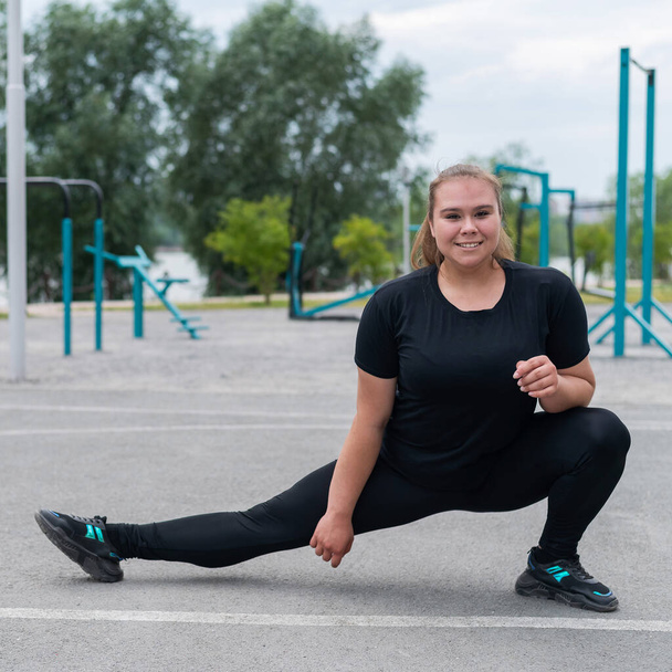 Obese young caucasian woman doing side lunges. Fat beautiful smiling girl in a black tracksuit is engaged in fitness outdoors. Warming up before training. - Φωτογραφία, εικόνα