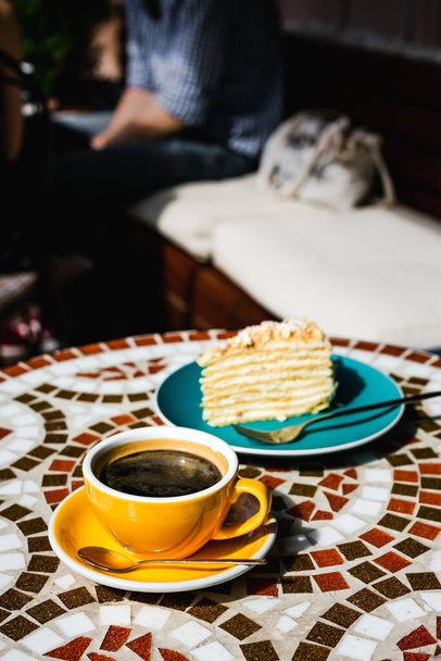 A slice of napoleon cake on a blue plate and a yellow cup of black coffee on a sunny day on a terrace of a cafe, mosaic stone table - Fotó, kép