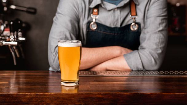 Reden Sie mit dem Barkeeper bei einem Drink. Mann in Schürze mit verschränkten Armen und leichtem Bier mit Schaum auf Holztheke - Foto, Bild