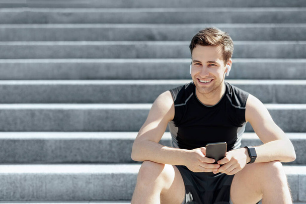 Glücklicher Typ mit Kopfhörer und Smartphone in der Hand, der sich vom Training erholt und auf der Treppe der Stadt sitzt - Foto, Bild