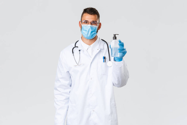 Covid-19, Gesundheitspersonal, Pandemie und Virusprävention. Schöner hispanischer Arzt in weißen Peelings, Arzt mit medizinischer Maske und Handschuhen, die Seife oder Händedesinfektionsmittel zeigen - Foto, Bild