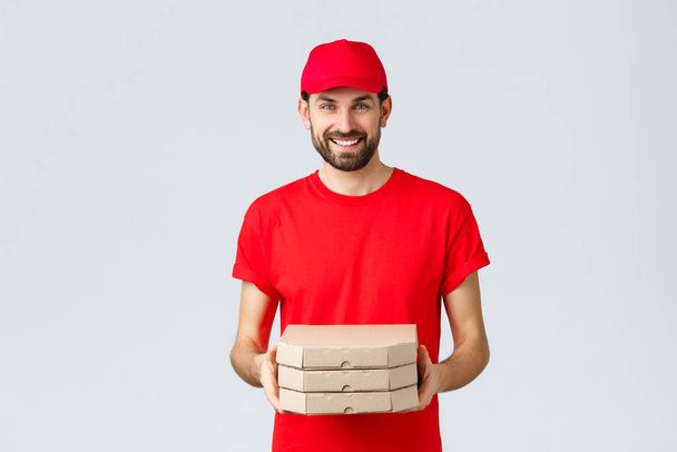 Food delivery, quarantine, stay home and order online concept. Friendly smiling courier in red uniform giving client order. Employee bring pizza at your house, standing grey background - Photo, Image