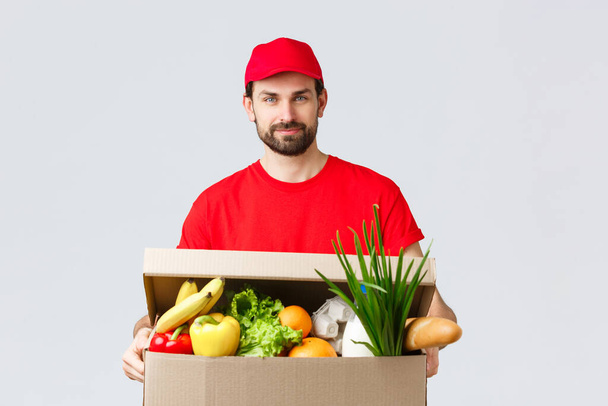 Groceries and packages delivery, covid-19, quarantine and shopping concept. Handsome courier in red uniform cap and t-shirt, bring grocery package, box delivery to client house, fast shipping - Foto, Bild
