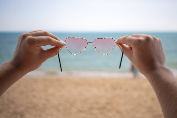 I love the sea. hands hold pink heart-shaped sunglasses. in the background a sandy beach and blue ocean in blur - Photo, Image