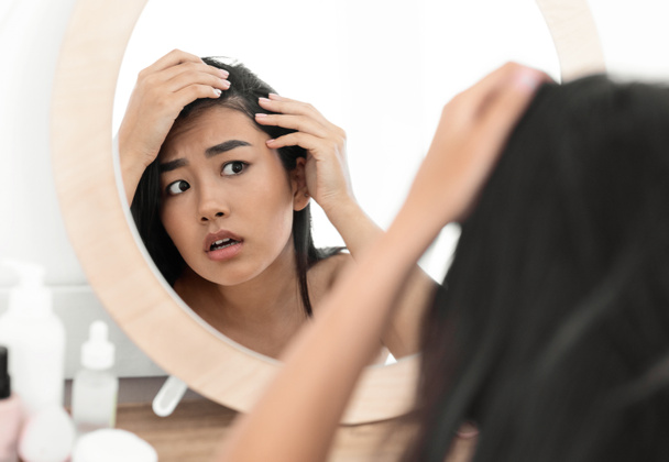 Беспокойная азиатка, проверяющая волосы в зеркале дома
 - Фото, изображение