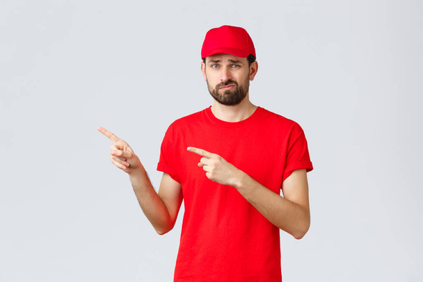 オンラインショッピング、隔離中の配達とテイクアウトの概念。赤のTシャツやキャップで指標と不満、消極的宅配便、失望し、アイデアのようにしない場合は、指を指して左に微笑んだ - 写真・画像
