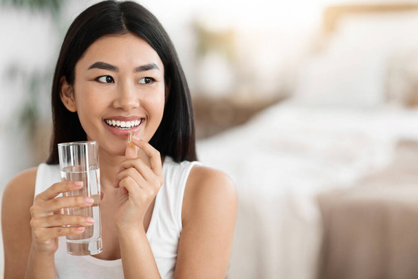 Ευτυχισμένη Ασιάτισσα που παίρνει χάπια και κρατάει ένα ποτήρι νερό. - Φωτογραφία, εικόνα