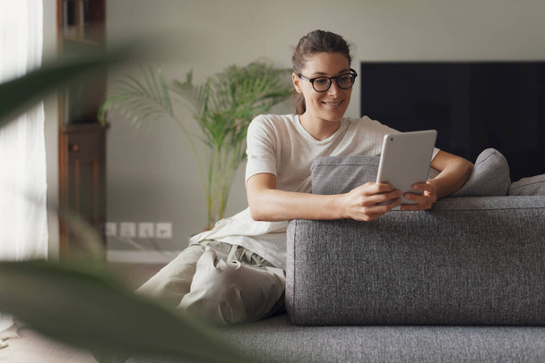 Νεαρή χαμογελαστή γυναίκα που χαλαρώνει στο σπίτι στο σαλόνι, κάθεται στον καναπέ και συνδέεται online με το ψηφιακό tablet της - Φωτογραφία, εικόνα
