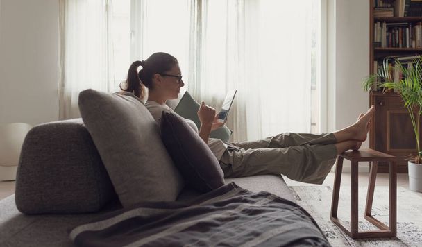 Woman relaxing on the sofa and connecting with her tablet in the living room - 写真・画像