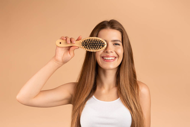 Portrait of pretty lady with long silky hair and wooden brush smiling on beige background - Fotoğraf, Görsel