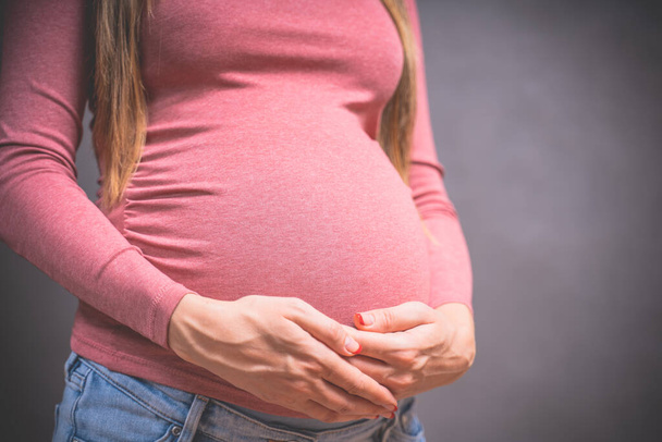 Έγκυος γυναίκα κρατά το χέρι στην κοιλιά σε ένα γκρι φόντο. Αντιγράψτε χώρο, εγκυμοσύνη, μητρότητα, τους ανθρώπους και την προσδοκία έννοια. Κοντινό πλάνο, αντίγραφο χώρου, σε εσωτερικούς χώρους. Γραμμένη φωτογραφία της εγκυμοσύνης. - Φωτογραφία, εικόνα