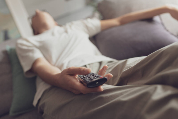Скучная женщина засыпает на диване, смотря телевизор, она держит пульт дистанционного управления
 - Фото, изображение
