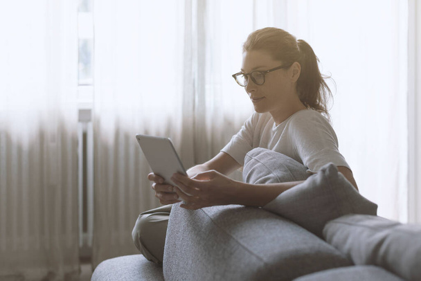 Młoda, atrakcyjna kobieta relaksująca się w domu w salonie, siedzi na kanapie i łączy się online ze swoim tabletem cyfrowym - Zdjęcie, obraz
