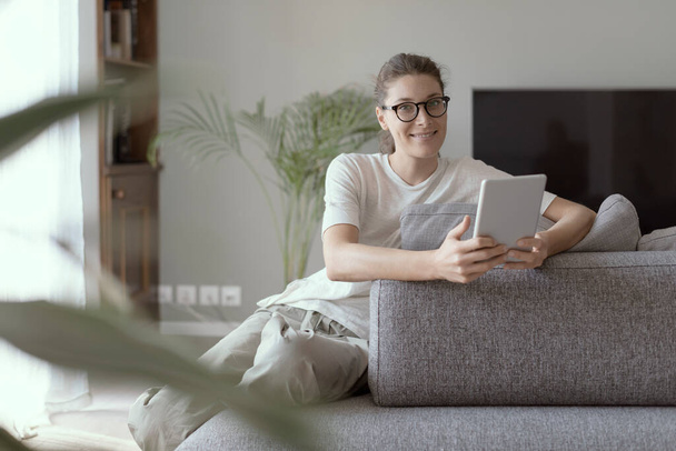 Young smiling woman relaxing at home in the living room, she is sitting on the couch and connecting online with her digital tablet - Φωτογραφία, εικόνα
