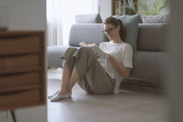 Glückliche Frau mit Brille sitzt zu Hause auf dem Boden und liest ein interessantes Buch - Foto, Bild