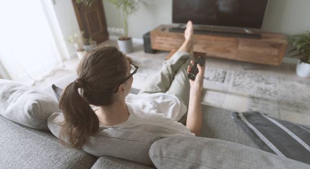 Young woman relaxing on the sofa at home and watching TV, she is holding the remote control - Fotó, kép