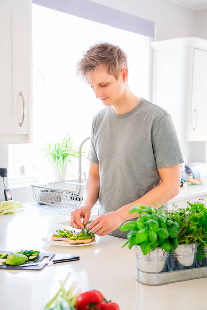 Young caucasian man making a healthy meal of avocado toast and vegetables putting just picked greenery from homegrown garden on the kitchen. Breakfast ideas. Vegetarian and vegan diet. Copy space. - Photo, image