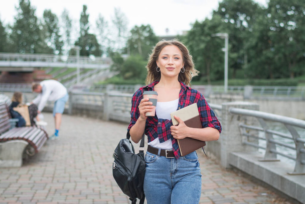 A beautiful young woman with long blond hair holding a coffee cup and texting on her smart phone while walking in a university campus a summer day - Valokuva, kuva