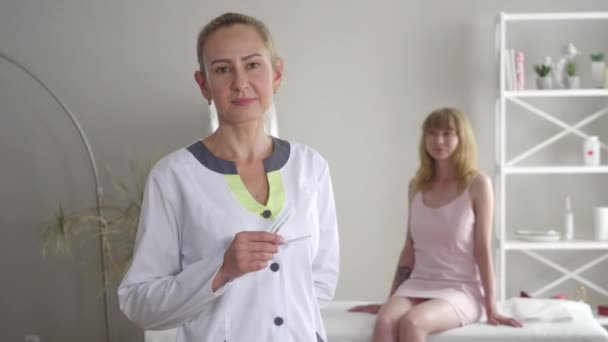 Portrait of confident acupuncturist posing with needles as young blond female patient sitting at the background. Adult Caucasian brown-eyed woman doing acupuncture in clinic. Occupation, lifestyle. - Footage, Video