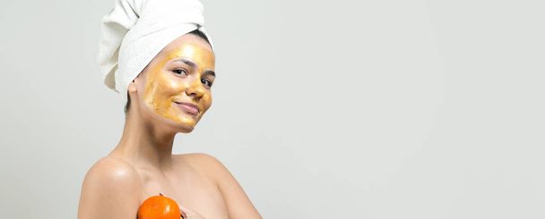 Schönheitsporträt einer Frau in weißem Handtuch auf dem Kopf mit goldener Pflegemaske im Gesicht. Reinigung der Haut Öko-Kosmetik Wellness-Entspannungskonzept. Ein Mädchen steht mit dem Rücken und hält eine orange Mandarine. - Foto, Bild