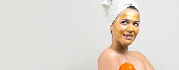 Schoonheidsportret van vrouw in witte handdoek op hoofd met gouden voedende masker op het gezicht. Huidverzorging eco biologische cosmetische spa ontspannen concept. Een meisje staat met haar rug en houdt een oranje mandarijn vast. - Foto, afbeelding