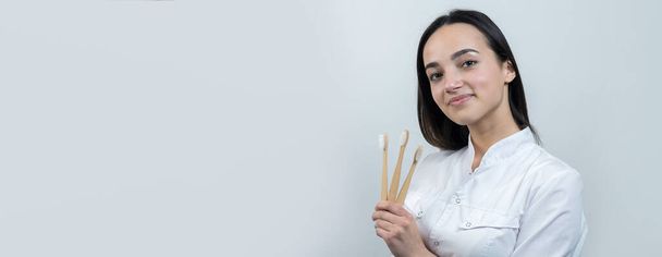 Una joven morena sostiene un cepillo de dientes ecológico en sus manos. Una chica con una bata blanca promueve cero residuos
. - Foto, Imagen