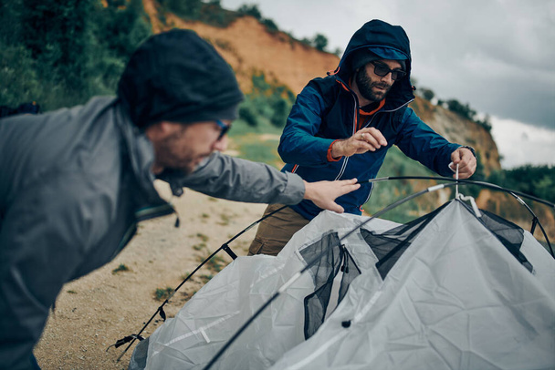 Dwóch atrakcyjnych mężczyzn demontujących namiot nad jeziorem na kempingu. - Zdjęcie, obraz
