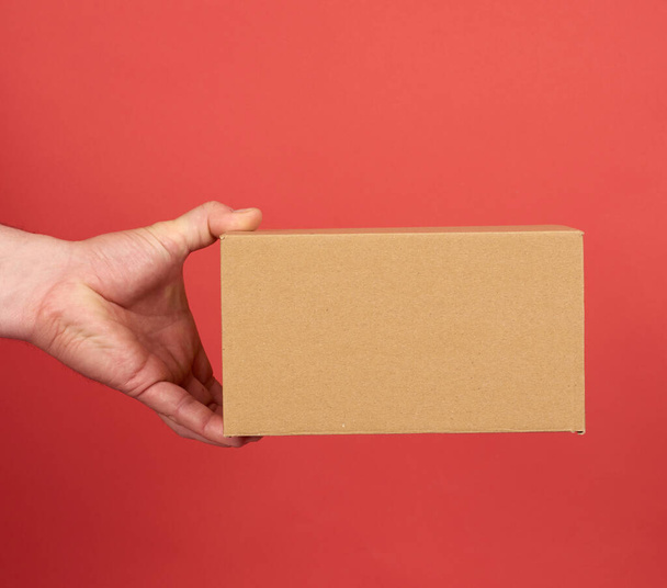 male hand holds a brown cardboard box on a red background, concept of goods delivery - Fotografie, Obrázek