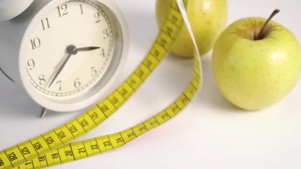 White analog alarm clock and green apples on a white table. A multi-colored bright centimeter descends from above. Slimming Sports Diet Concept - Footage, Video