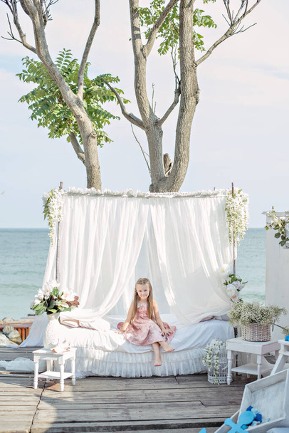 Delicate little girl blonde with long hair in a beautiful pink powdery dress sits by a white bed with chiffon canopies, decorated with wisteria, flowers against the background of the sea. Design in a marine style. - Φωτογραφία, εικόνα