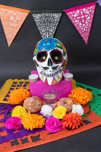 Traditional Mexican Day of the dead offering altar with cut paper, a hand painted skull, bread, flowers, candles and mezcal on black background - Photo, Image