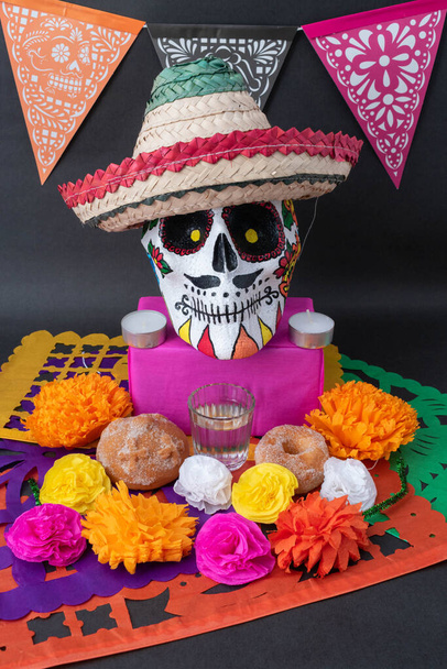 Mexican Day of the dead altar with traditional bread, colorful flowers, banner, a hand painted skull with a hat and cut paper on black  - Photo, Image